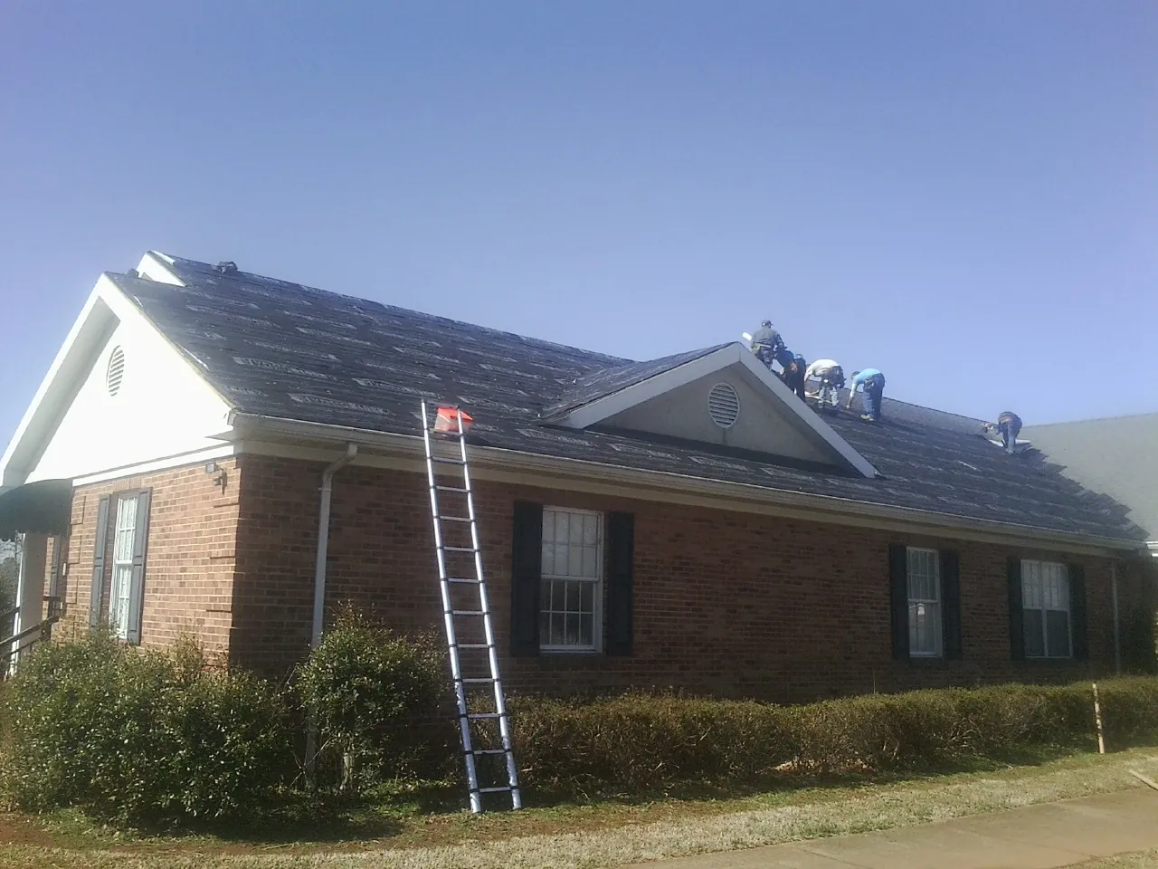 Crew tearing shingles off a brick ranch roof with a ladder set — Metal Roof Repair work by our crew in Eagle Bay