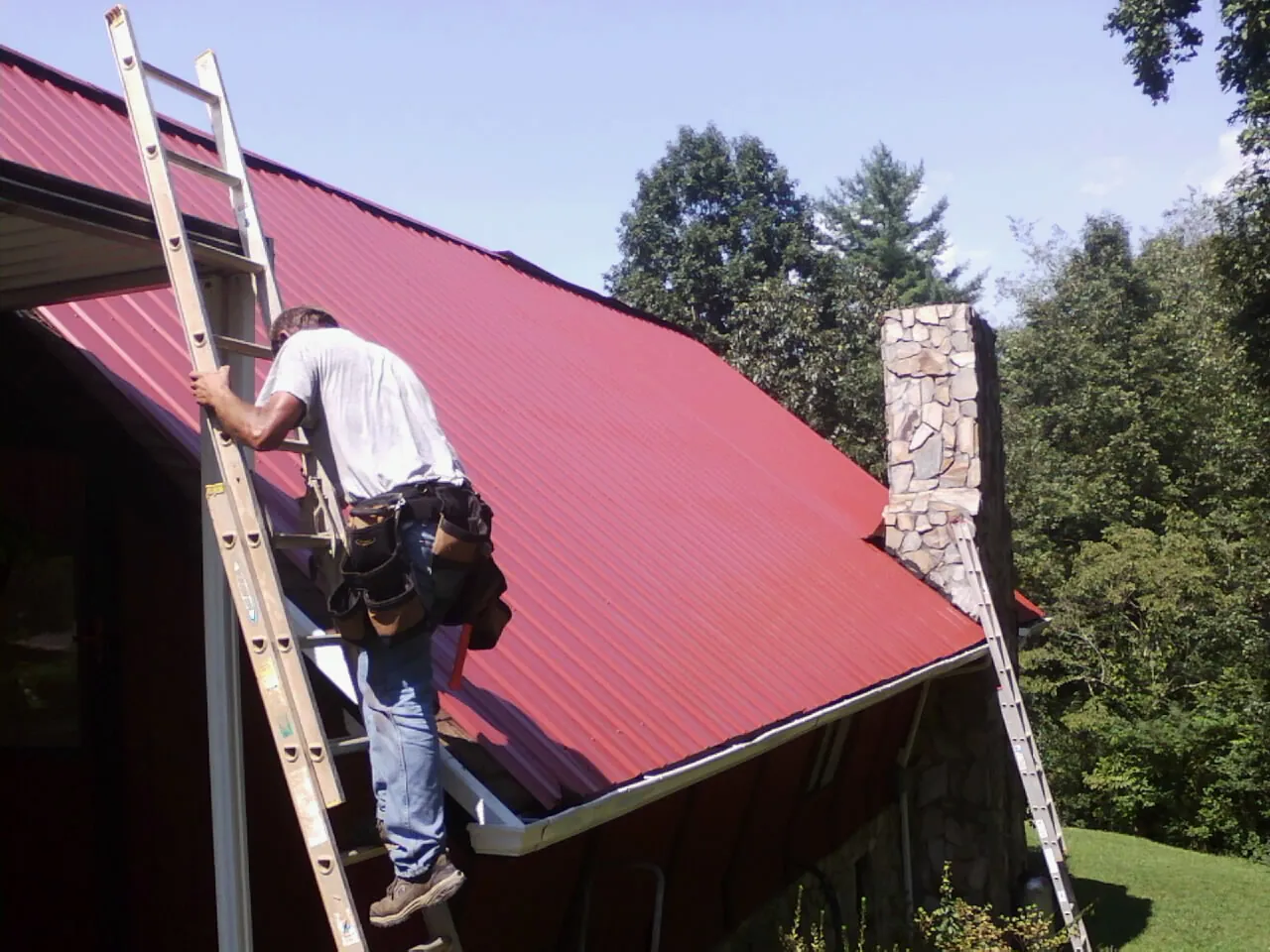 Roofer on a ladder beside a red metal roof and stone chimney — Soffit & Fascia Repair by Roofing Brothers in Eagle Bay